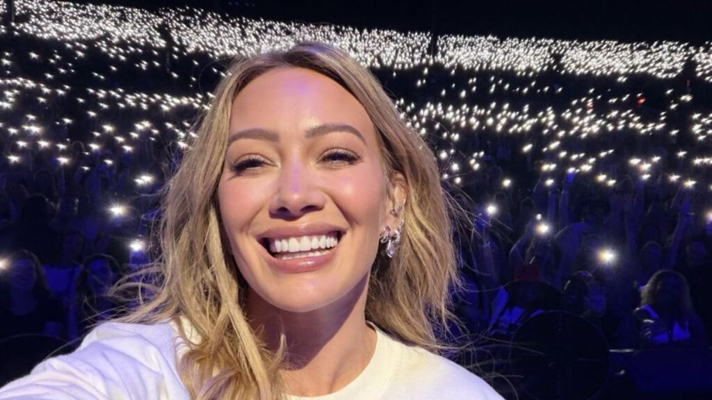 Hilary Duff durante apresentação ao vivo, com fãs animados no fundo